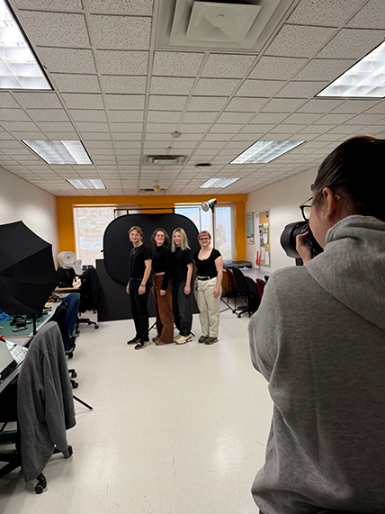 Photographer taking a group portrait of four people standing in front of a black backdrop in a brightly lit room with photography equipment.