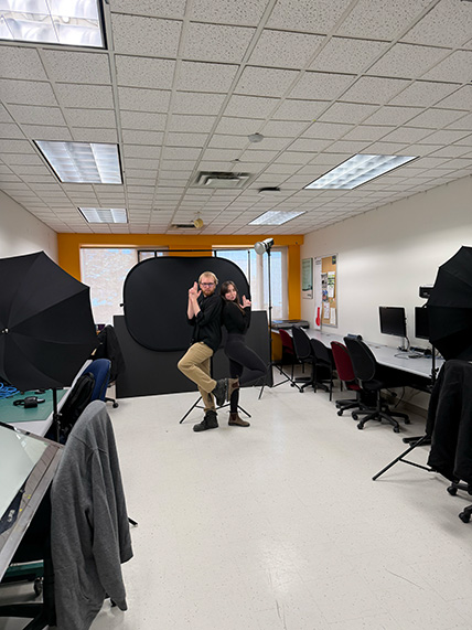 Two people posing back-to-back with finger guns in a photography studio with lighting umbrellas and computer workstations.