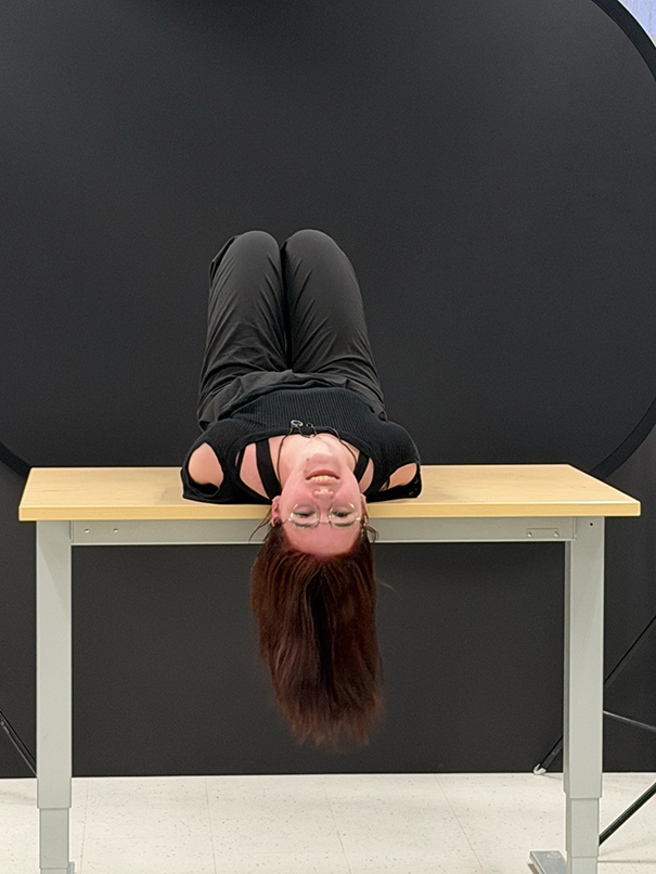 Person wearing glasses and black clothes lying upside down on a wooden table against a dark background.