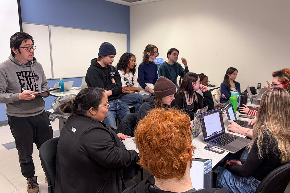 A diverse group of people in a classroom meeting around a table, some working on laptops and tablets while one person stands holding a tablet.