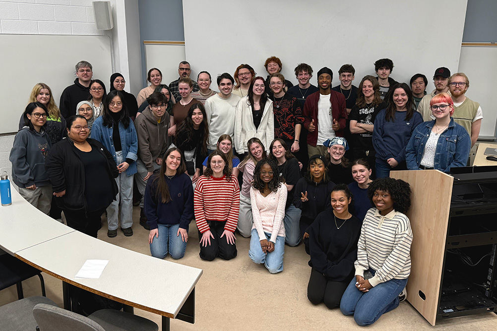 Large group of smiling students posing together in a classroom with tiered desks and a podium.