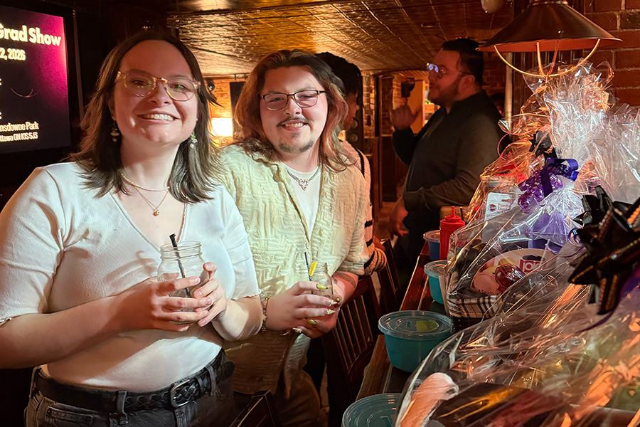 Two people smiling and holding drinks, standing next to a bar decorated with gift baskets wrapped in clear plastic.