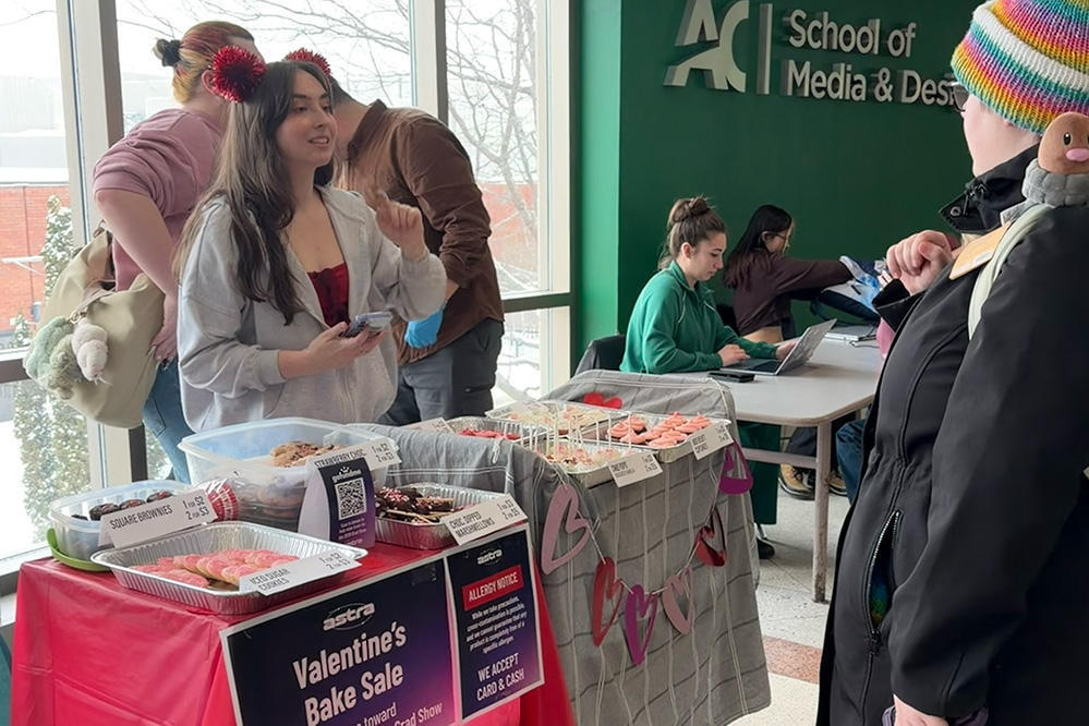 People at a Valentine's Bake Sale table with various baked goods displayed and a sign for Astra at the School of Media & Design.